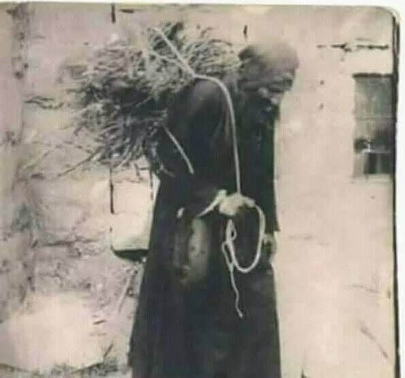 Vintage photograph of Saint Charbel in his monastic habit carrying a large bundle of firewood on his back outside a stone wall